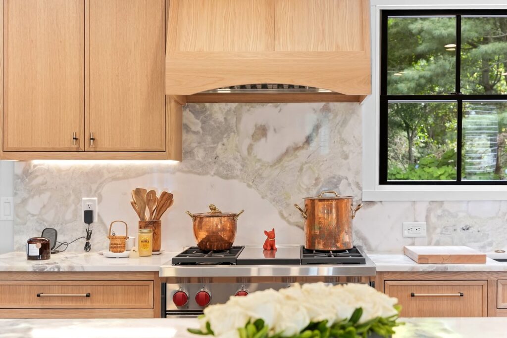 A bright kitchen with wood cabinets, a marble backsplash, a stove with pots, and a window overlooking greenery.