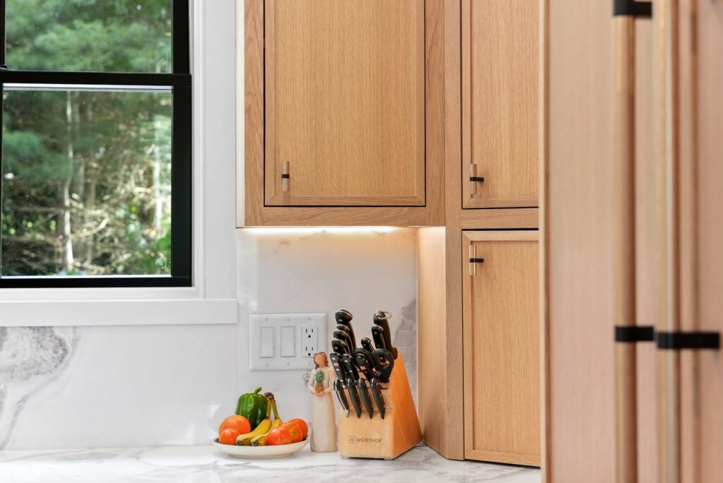 A bright kitchen corner with wooden cabinets, a window, and a knife block beside fresh vegetables.