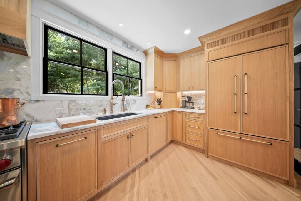 Bright kitchen with light wood cabinets, marble countertops, stainless steel appliances, and a large black-framed window.